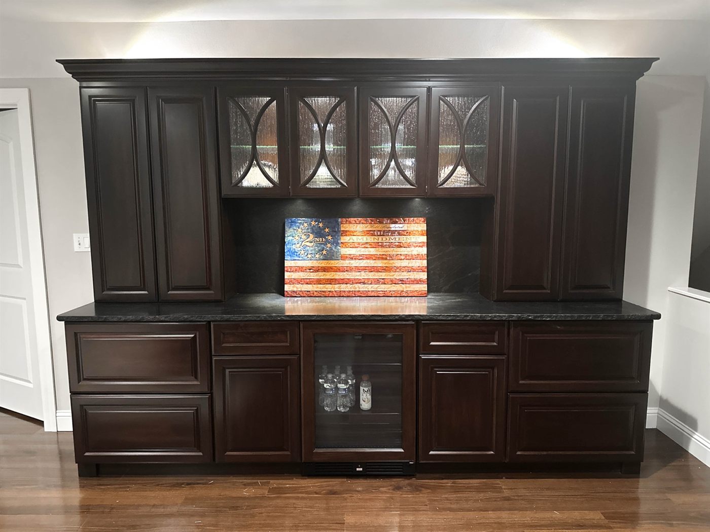 Built-in cabinetry with lit glass upper doors, LED-washed backsplash, granite top and a beverage fridge below
